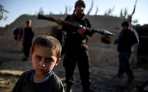 A young boy looks at the camera as a policeman holding a rocket-propelled grenade stands behind in a house at Deh Qubad village in Maiwand district of Kandahar province on September 27, 2020.