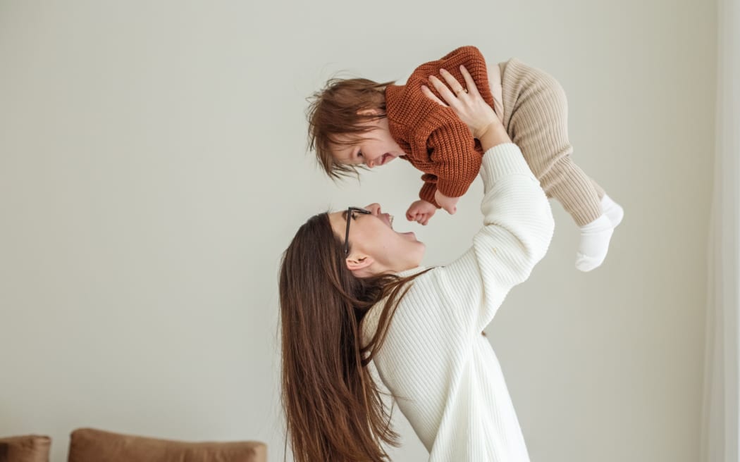 Happy cheerful young mother throws an adorable baby in air, plays, hugs baby with love, care, enjoys motherhood, maternity leave. Family spends time together.