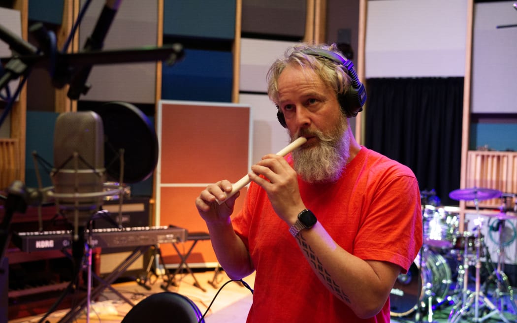 A man with a beard wearing a red t-shirt and headphones playing a traditional flute.