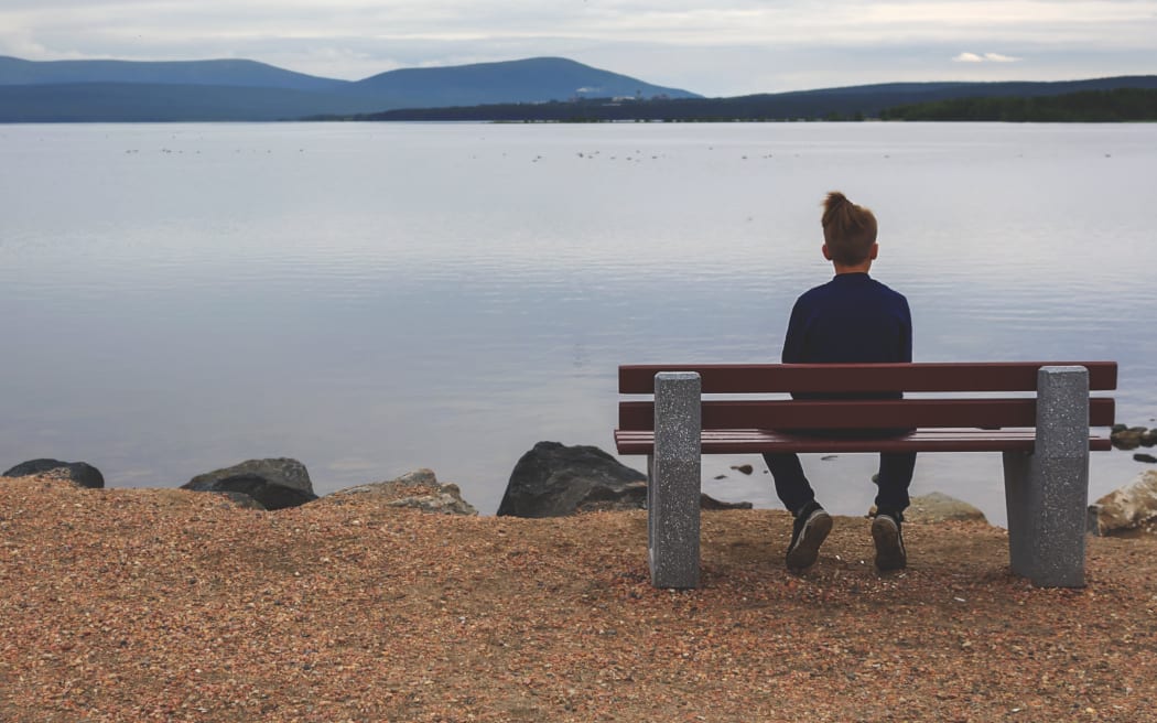 Teenager sitting alone on bench on lake in the evening, looking at horizon and thinking about future. Unity with nature. Loneliness of youth. Psychological assistance to young people.