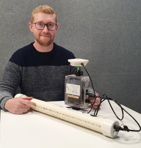 A photo of Ben McEwen seated at a table with his audio lure system. It's about 30 centimetres high and consists of a thermal camera in a protective casing with a microphone mounted on top. Also pictured is a long reuseable battery that charges the device.