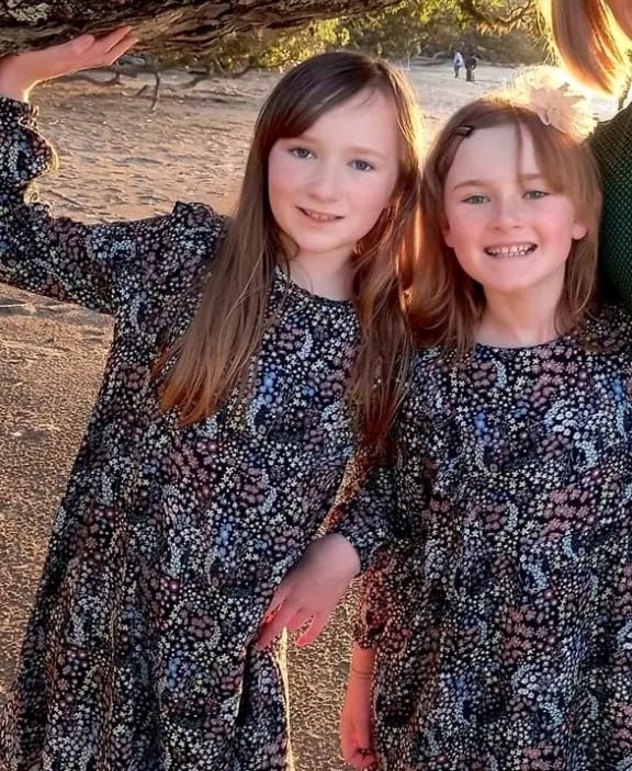 Two sisters, probably Pākehā, aged 8 and 10 in matching navy-blue frocks with a busy floral pattern, smiling at camera. One has long brown hair the other possibly shoulder-length or longer but hidden behind her back.