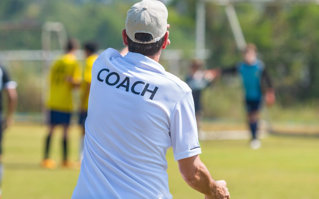 Back view of male football coach in white COACH shirt at an outdoor football field giving direction to his football team