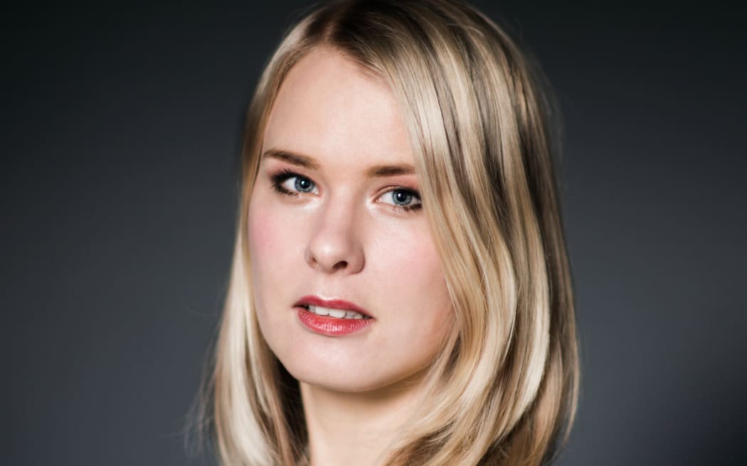 Emma Vitz looks at the camera with a serious expression. She is pictured from the shoulders up, wearing a white blouse and standing side on to the camera. She is against a dark backdrop.