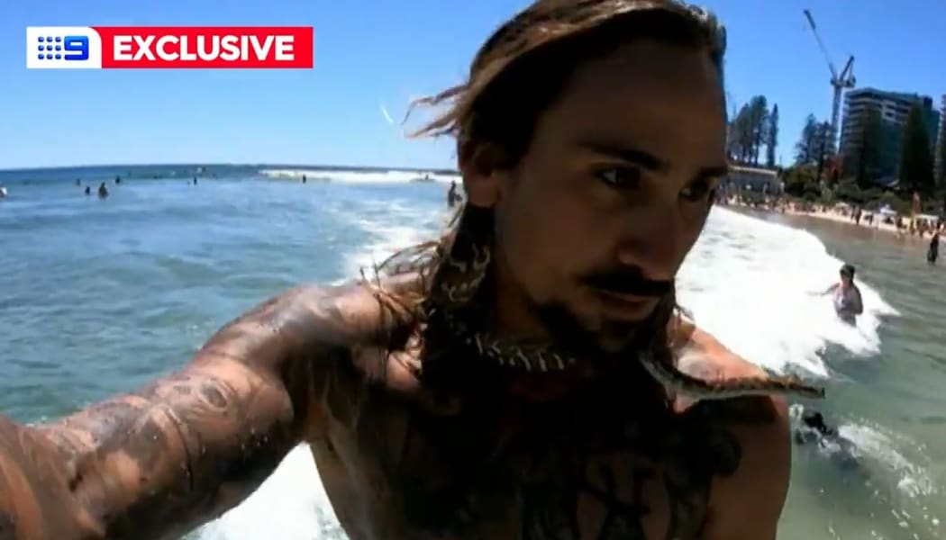 A screenshot of a 9News piece about Gold Coast man Higor Fiuza surfing with his bredli carpet python Shiva around his neck.
