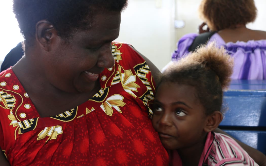 Une femme en robe rouge tient une petite fille dans une étreinte.  Ils se sourient.