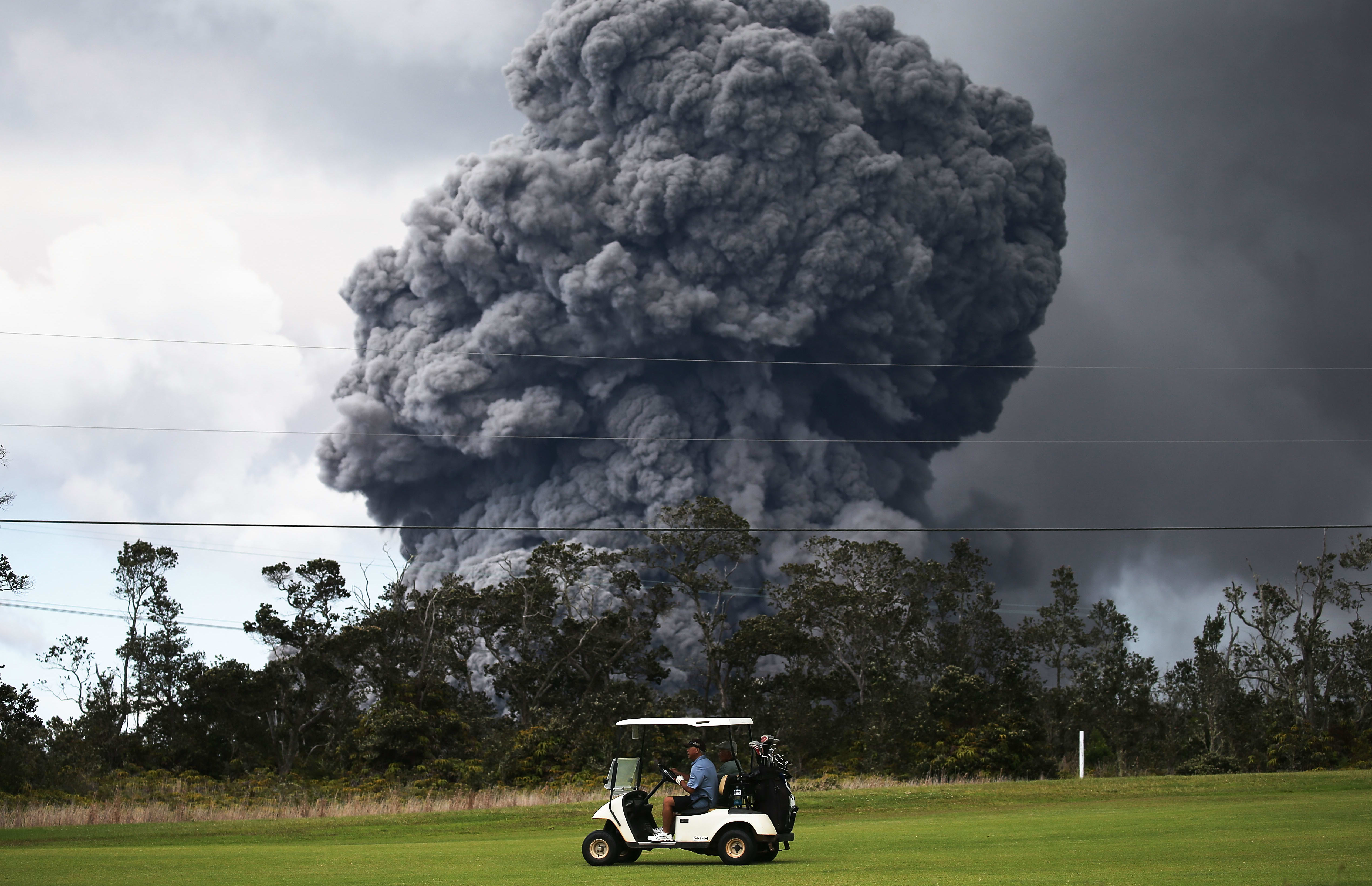 A man drives a golf cart at a golf course as an ash plume rises in the distance from the Kilauea volcano on Hawaii's Big Island on May 15, 2018