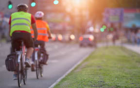 Two cyclists with protective equipment are approaching an intersection in a busy part of the city towards the setting sun.
