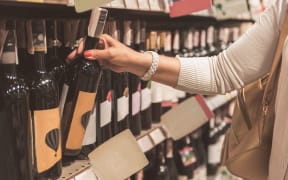 Woman is standing near shelf with bottle of wine and choosing one. Close up of female hand, rising beverage
