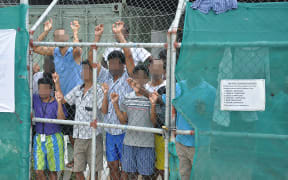 Asylum seekers behind behind a fence on which a sign orders guards to carry the hooked knives used to cut the rope of people who attempt to hang themselves.