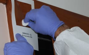 A close up photo of two hands with gloves on, swabbing within a 100 square centimetre stencil stuck to a wall. By swabbing within that area, testing companies get a sample that corresponds to Ministry of Health guidelines for meth.
