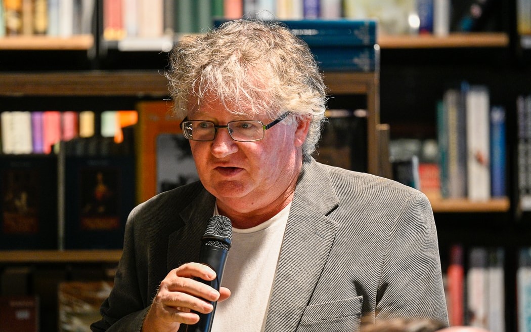 Portrait of Russell Kirkpatrick wearing glasses and speaking into a microphone in front of book shelves.