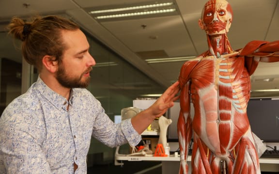 A man in a shirt sits next to a model of the human body with muscles visible. He is touching the bicep of the model.