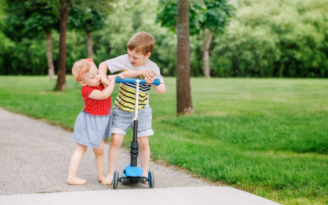 A photo of two little Caucasian preschool children fighting hitting each other. Boy and girl can not share one scooter. Older sibling brother not giving his toy to younger sister. Communication problems.