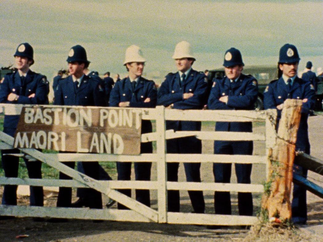 A sign says "Bastion Point Maori Land" 6 police officers are standing behind the fence and sign.