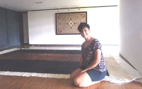 Ruha Fifita sitting in front of her enormous tapa, with another tapa by Christelle Montané on the wall.