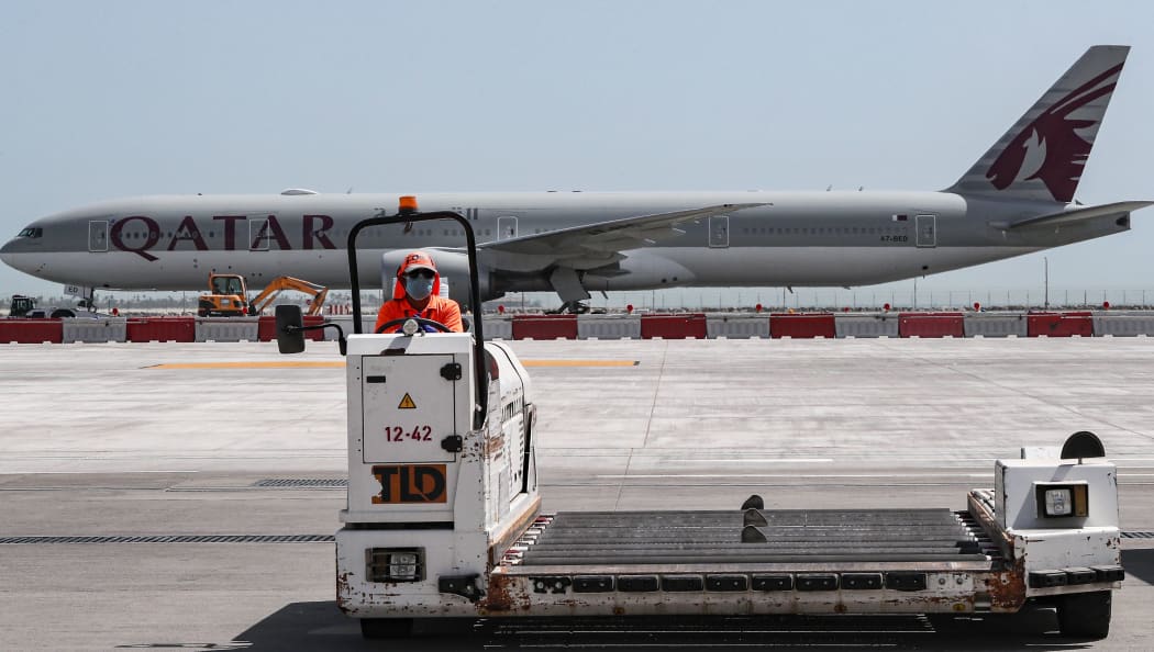 (FILES) In this file photo taken on April 1, 2020, an airport worker wearing a face mask (protective measure during the COVID-19 coronavirus pandemic) mans a luggage trolley while behind him is seen a Qatar Airways Boeing 777 aircraft at Hamad International Airport in the Qatari capital Doha.