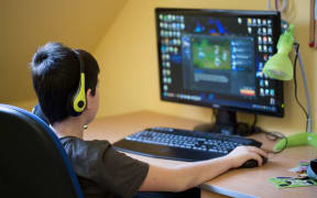 A photo of a teenager boy taken from behind. He is using computer at home with headphones to play a game in his room.
