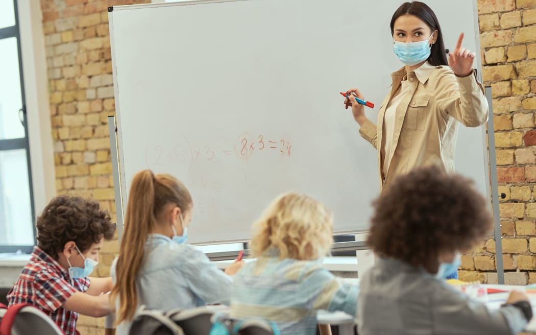 Female teacher wearing protective mask during coronavirus pandemic writing mathematic task on board for group of elementary school kids. Education, math and covid19 concept