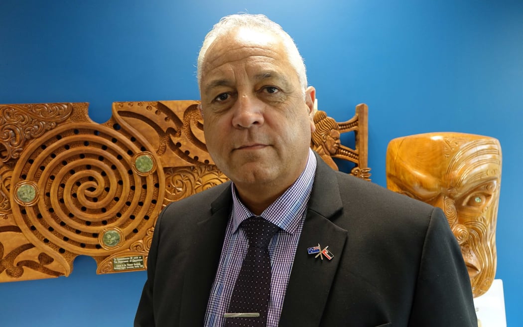 Head and shoulders shot of man infront of carved wooden Maori wall panel and carved  head.