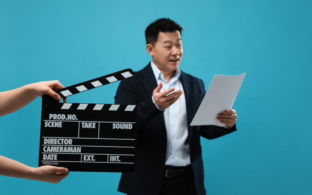 Happy asian actor performing role while second assistant camera holding clapperboard on light blue background, selective focus. Film industry