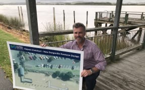 Kaipara District Council Mayor Jason Smith at Kaipara Harbour's Dargaville wharf with Dargaville pontoon rebuild design outline, the pontoon to be rebuilt where piles can be seen in photo background.