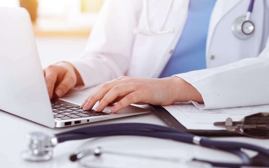 Unknown woman-doctor typing on laptop computer while sitting at the table in sunny clinic.