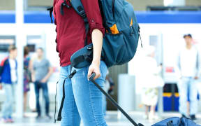 A woman with a backpacker wheels a suitcase at an airport