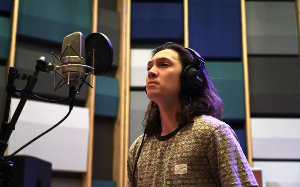A man with long hair wearing a t-shirt and headphones stands in front of a microphone.