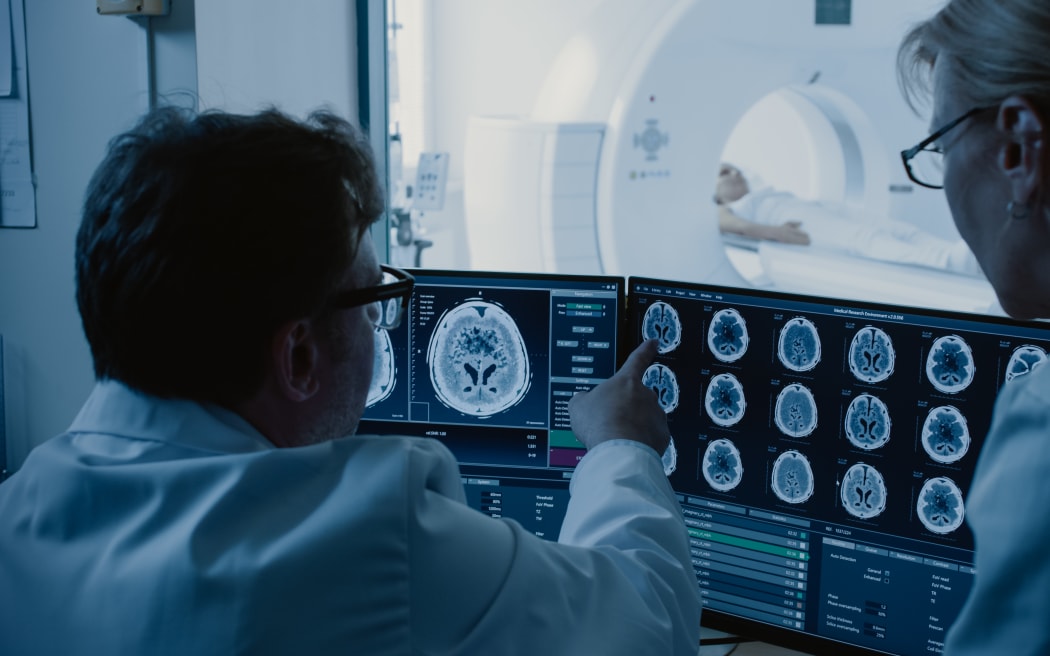 In the control room, the doctor and radiologist discuss the diagnosis while watching the procedure and monitors displaying brain scan results, and in the background the patient undergoes an MRI or CT scan procedure.