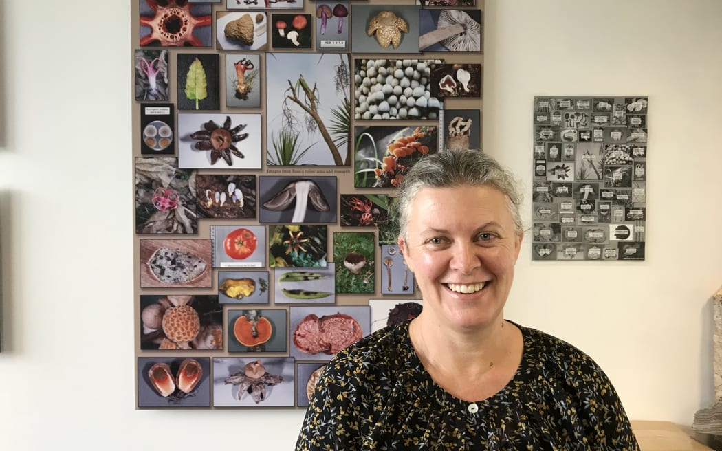 Megan stands in front of a collection of images of different fungi in the foyer area of Manaaki Whenua collection area. She is wearing a black, buttoned blouse and smiling.