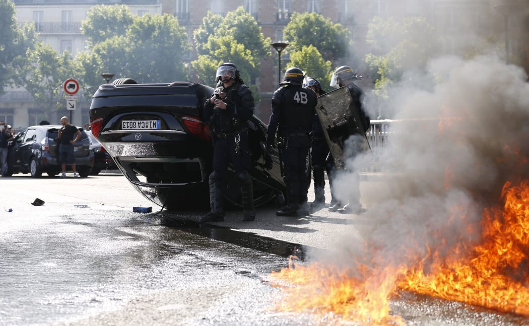 Smoke rises from a fire burning next to French CRS riot police standing near an overturned car as taxi drivers block Porte Maillot in Paris.